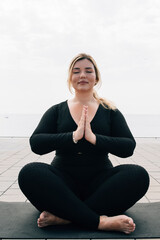 Fototapeta na wymiar beautiful blonde plus size girl meditating in front of the sea with a mat and dressed in sports clothes