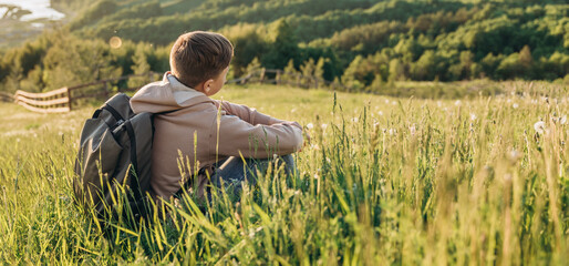 Tourist with backpack sitting on top of hill in grass field and enjoying beautiful landscape view....