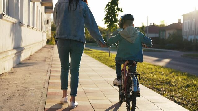 Happy family walk on street the city mother teaches her son child to ride a bike and run nearby. Slow motion woman helps keep balance for safe activity. Sports walk in nature. Healthy lifestyle.