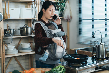 busy asian new mother is having a phone talk while frying food for breakfast by the kitchen stove with her baby daughter in the carrier at home. - Powered by Adobe
