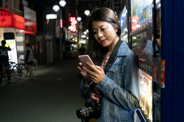 pretty asian woman tourist standing by a vending machine and using cellphone to look for directions on a shopping street in Osaka city in japan in the evening - obrazy, fototapety, plakaty