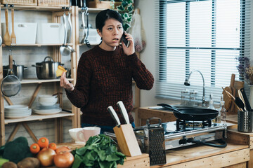 unhappy asian woman eating bread is complaining with hand gestures while talking on the phone by the kitchen stove at home in the morning.