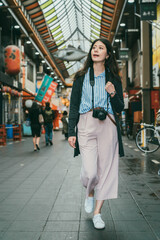 full length with vertical shot of relaxed asian female traveler with camera looking into distance while exploring inside local kuromon ichiba market in Osaka japan