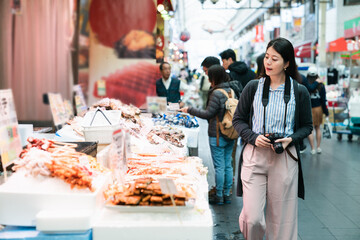 asian woman traveler with camera passing by a local stall selling Japanese processed seafood while visiting kuromon ichiba market in Osaka japan