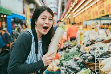 portrait of excited asian female tourist holding white radish and looking at camera with her mouth open at local stall selling fresh produce in kuromon ichiba market in Osaka japan