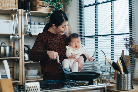 Multitasking Pretty Asian Mother Is Smiling And Talking To Her Adorable Baby In Arm While Making Breakfast With A Spatula And Frying By The Stove In The Kitchen