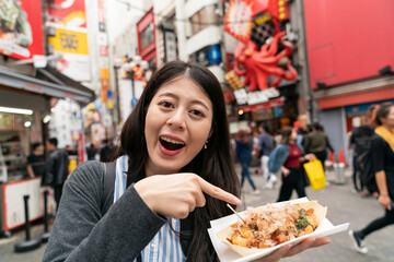 excited Asian girl sight-seer smiling at camera and finger pointing at Japanese snack takoyaki octopus balls while shopping in shinsaibashisuji and doutonbori Osaka japan
