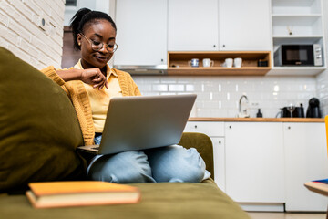 Young African American woman sitting on sofa at home studying using laptop.	
