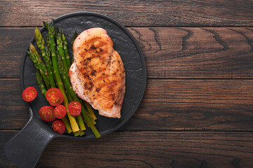 Chicken breast grilled with vegetables. Grilled chicken steak, asparagus and cherry tomatoes in black plate on wooden dark table background. Healthy diet lunch. Barbecue steak fried. Top view.