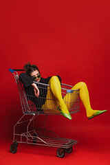 Funny portrait of stylish young girl in yellow tights sitting on shopping trolley, posing isolated...