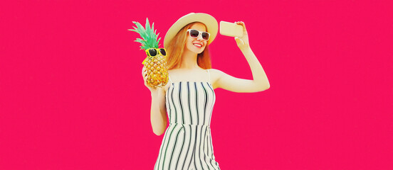 Summer portrait of happy smiling woman with pineapple taking selfie by smartphone wearing straw hat, striped jumpsuit, sunglasses on pink background
