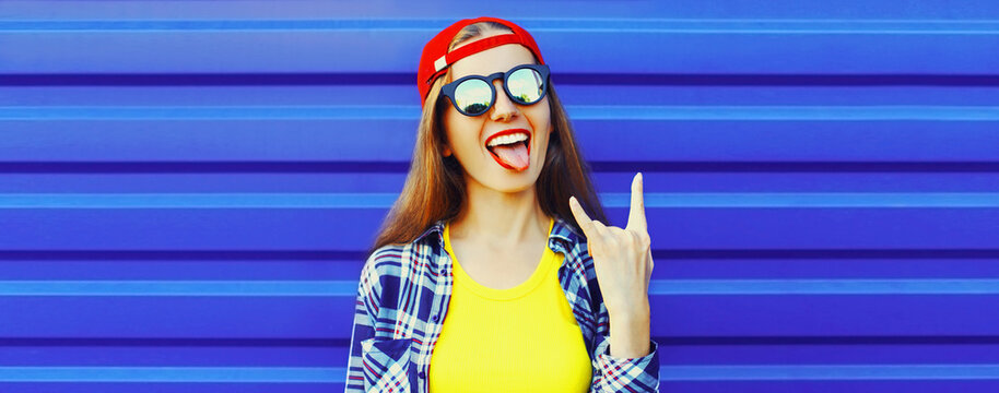 Portrait of happy cheerful young woman showing her tongue having fun wearing red baseball cap on blue background