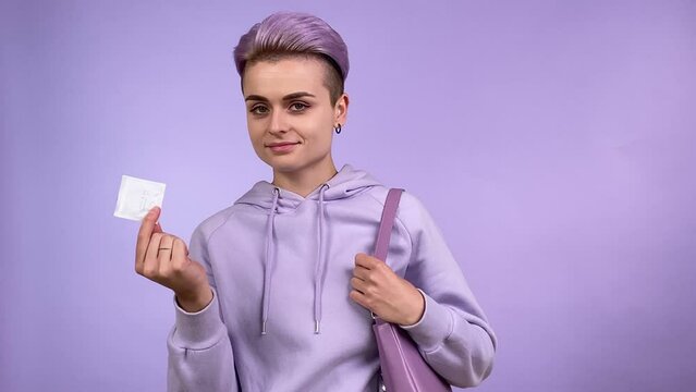 Positive young adult woman with short dyed hair holding showing condom, putting contraceptive device in handbag, looking at camera, isolated on purple background indoors. Monochrome concept