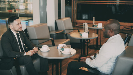 Two multi-ethnic businessmen sitting, smiling and discussing their startup in airy cafe during lunch time. Coffee cups are on their table