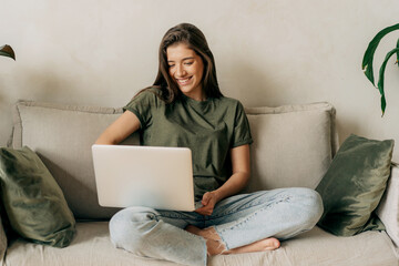 Young happy smiling latin female student sitting on sofa uses laptop for online learning.