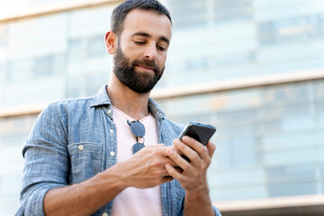 Pensive bearded latin man holding smartphone chatting, check email, reading news outdoors. Handsome hipster using mobile app shopping online on the street. Mobile banking concept 