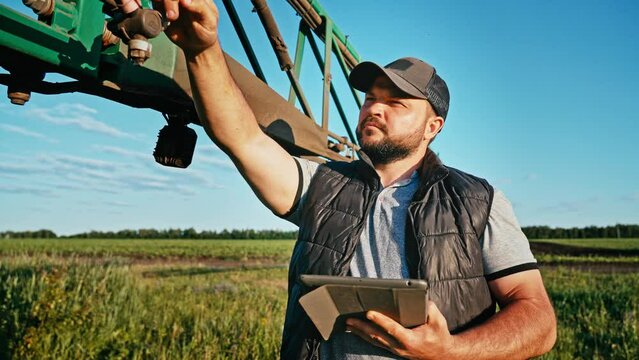 Male farmer engineer stand next to tractor and uses mobile application on digital tablet PC. Mechanic online optimizes work of small businesses in production of products and repairs machinery.