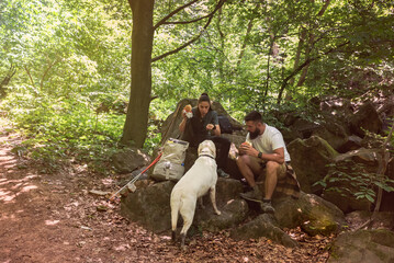 Fototapeta premium Young hikers couple sitting on the rocks on the mountain woods eating a sandwiches at break while their dog watch them. Nature lovers hiking through the forest eating lunch. With film grain