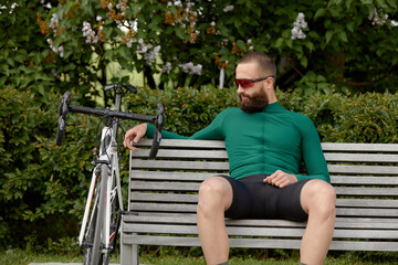 Fototapeta premium Stylish handsome guy cyclist rests on a wooden bench near bicycle in summer park.