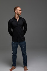 An attractive white man in jeans and black shirts tands in full growth against a gray background. Copy space