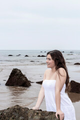 Fototapeta na wymiar young brunette woman leaning on a rock on the beach in a white dress
