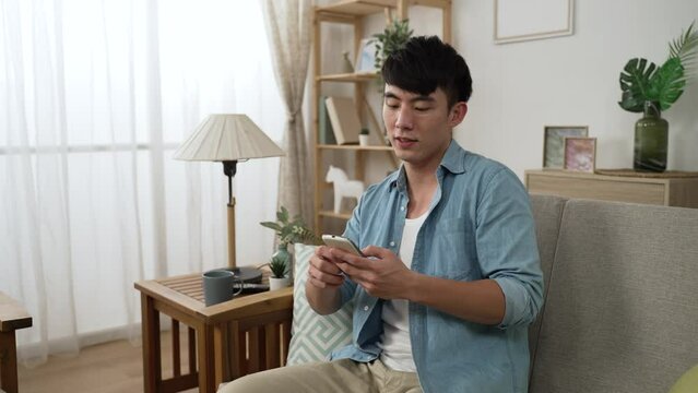 leisure Chinese male approaching and sitting down on the comfortable living room sofa to enjoy social media feeds on his smartphone at home during daytime.