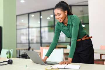 Wholly directed on task young african-american female employee stands near desk and using modern...