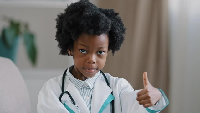 Close-up little cute kid girl in medical gown looking at camera posing indoors smiling pretending be doctor plays nurse showing thumb up gesture approval promotes dream job future profession concept