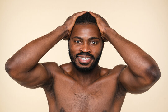 Sexy naked shirtless african-american young man looking at camera showing muscles looking at camera, taking care of skin, male beauty isolated in beige background