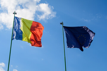 European Union and The Romanian national flags blowing in the wind towards cloudy blue sky in a sunny sprng day.