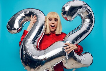 a happy, enthusiastic woman in a red shirt stands on a blue background and holds inflatable balloons in the shape of the number twenty-two in silver color. Horizontal Studio Photography