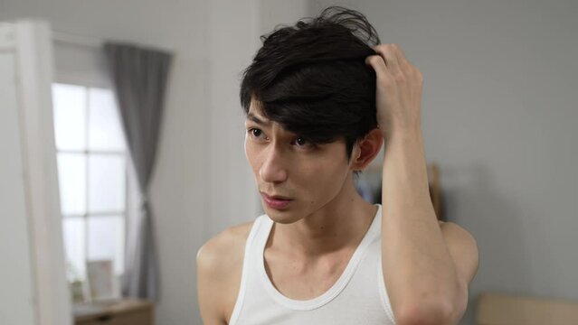 cheerful asian man in undershirt is looking into the mirror and arranging his hair during morning beauty routine in the bedroom at home.