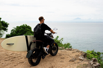 Fototapeta premium young man sits on a motorcycle with a surfboard.