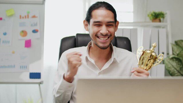 Confidential happy young Asian businessman celebrate with award, talking on VDO conference online and holding cup of successful winner trophy, cheerful man success and get prize in business