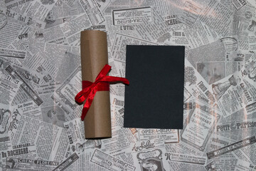 wooden board with black copy space on a newspaper background, next to the board is a scroll with a red bow, creative invitation, greeting card