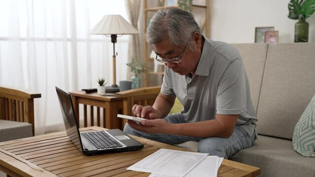 Retirement financial planning concept. senior asian man counting on calculator with family budget sitting in living room sofa using laptop computer and paper indoors