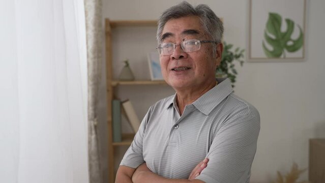 cheerful senior asian man looking at camera and smiling. confident old male enjoy retire life at home alone. happy grandpa looking in lens with hands and arms cross indoors