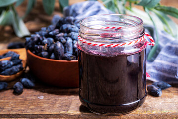 Small glass jar with homemade  haskap berry jam, on wooden background with bowl of fresh haskap...