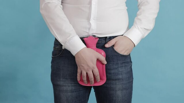 A man puts a red heating pad with hot water in the groin area. The concept of treatment of male diseases, prostatitis and bladder diseases, close-up