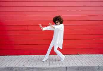 Afro woman with sunglasses gesturing on sidewalk in front of red wall