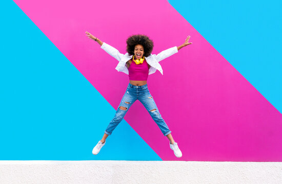 Playful woman jumping with arms outstretched in front of pink and blue wall