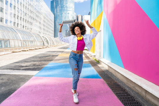 Happy young Afro woman with hands raised walking by colorful wall