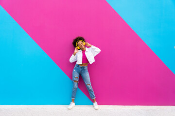 Young woman enjoying music listening through wireless headphones in front of pink and blue wall