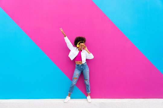 Happy Afro woman with hand raised enjoying music dancing in front of pink and blue wall