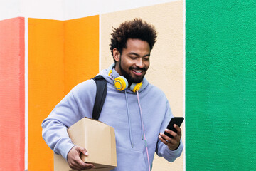 Smiling delivery man holding box using smart phone in front of colorful wall