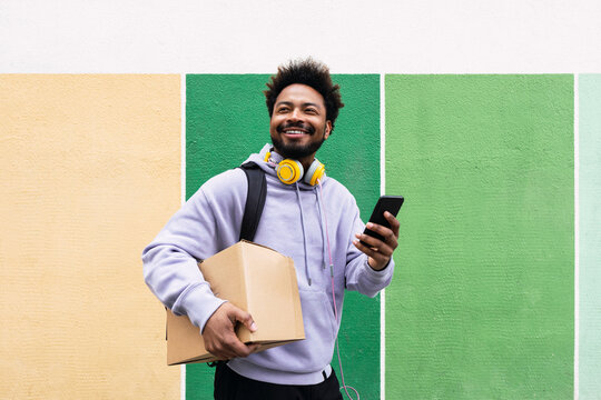 Smiling delivery man holding box and mobile phone in front of colorful wall