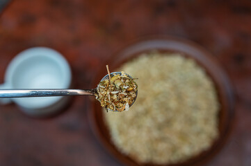 Full spoonful of dried Matricaria chamomilla flowers above the kitchen table. Brown bowl of medicinal herbs blurred in the background. Process of making an herbal tea or infusion of chamomile.