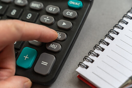Close-up of male hand doing calculations on calculator. Index finger presses the percentage button. An open spring-loaded notebook and a pencil lie side by side on a gray surface. Selective focus.
