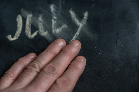 The semi-erased word JULY on the black chalkboard. An adult man's left hand removes the handwritten word with his fingers. Fuzzy letters on black surface. End of calendar month concept - Powered by Adobe