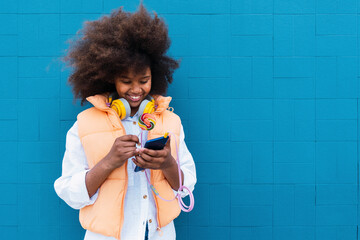 Smiling girl holding colorful lollipop using smart phone in front of blue wall